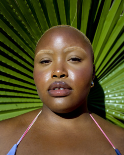 Close-up portrait of a Black woman with a shaved head and fair eyebrows, posing in front of a large green palm leaf.