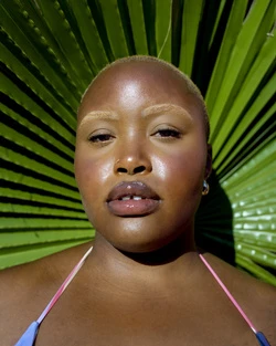 Close-up portrait of a Black woman with a shaved head and fair eyebrows, posing in front of a large green palm leaf.