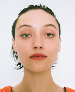 Headshot of a young woman with wet hair looking directly at the camera, wearing an orange top against a white backdrop.
