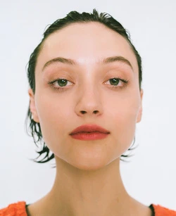 Headshot of a young woman with wet hair looking directly at the camera, wearing an orange top against a white backdrop.