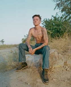 Shirtless person with tattoos and denim jeans sitting on a rock outdoors captured by Maiwenn Raoult