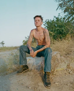 Shirtless person with tattoos and denim jeans sitting on a rock outdoors captured by Maiwenn Raoult