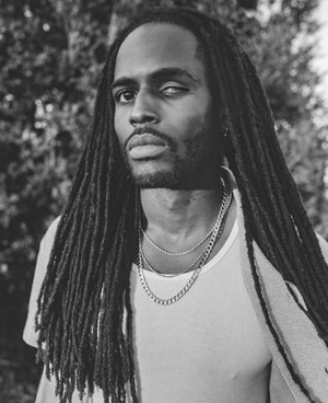 Monochrome portrait of a Black man with long dreadlocks looking at the camera wearing layered silver necklaces with a partial green background.
