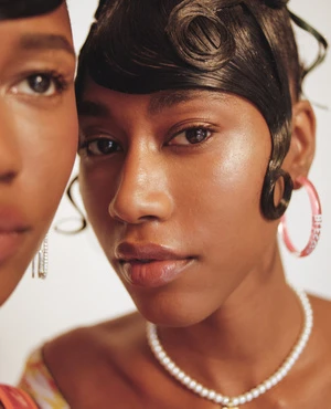 Close-up portrait of two black women with dark, styled hair, pearl necklace, and hoop earrings against a bright background.