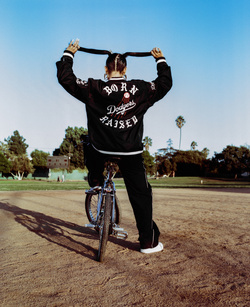 Woman with pigtails rides a blue bicycle on a dirt path with grass and palm trees in the background, wearing a jacket with text.
