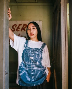A young woman with long dark hair stands in a doorway wearing a white T-shirt with denim overalls print and black pants in soft lighting.