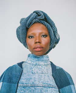 Close-up portrait of an adult female with dark skin, wearing a blue patterned head wrap and matching denim top and jacket, against a grey background.