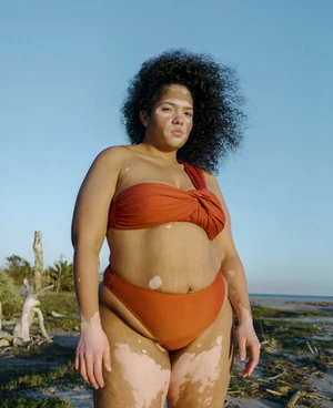 A curvy woman with vitiligo poses confidently in a rust-colored swimsuit on a sandy beach with a clear blue sky in the background.