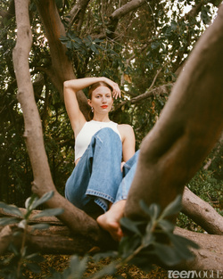 Woman in white crop top and blue jeans seated in a large tree by Maiwenn Raoult