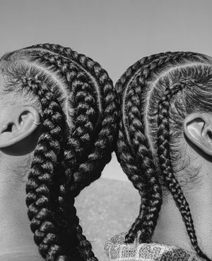 Close-up portrait of two individuals with identical braided hairstyles, set against a light gray backdrop, emphasizing hair texture and symmetry.