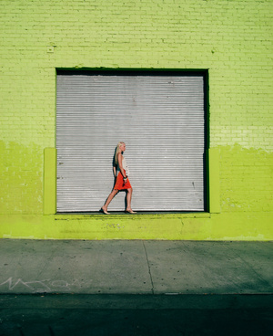 A lone blonde woman in an orange dress walks across a gray sidewalk, next to a green painted brick wall with a large, closed metal roll-up door.