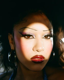 Close-up portrait of a woman with dramatic eye makeup, glossy red lips, and hoop earrings, partially shadowed on a light background.
