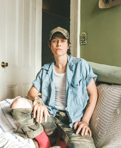 A woman with short brown hair, wearing a baseball cap, button-down shirt, and camouflage pants, sits on a couch in an indoor setting.