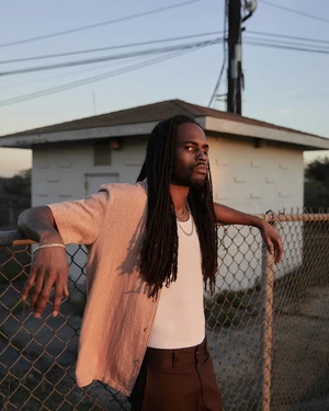 Black man with long dreadlocks leans against a wire fence outside in the golden hour with a neutral expression and casual clothing.