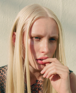 Portrait of a blonde woman with long hair and light eyebrows posing against a textured wall in Los Angeles.