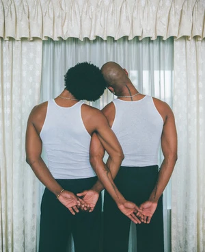 Two men in white tank tops and dark trousers standing back to back before a window with sheer curtains.