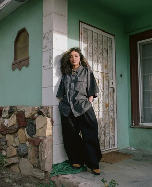 A young woman with curly hair in a relaxed pose against an exterior wall, wearing a dark denim shirt and baggy pants in a natural light setting.