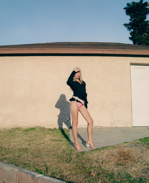 A slender woman poses in a black jacket and pink bikini bottoms in front of a beige stucco wall with green grass and a blue sky.