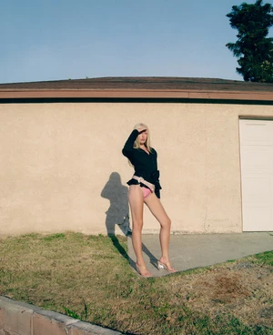 A slender woman poses in a black jacket and pink bikini bottoms in front of a beige stucco wall with green grass and a blue sky.