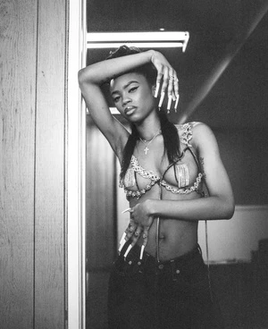 Monochrome portrait of a young adult woman in a beaded top and jeans leaning against a wall with decorative nails and jewelry indoors.