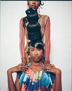 Two African-American women pose in a photo studio; the woman in front wears a colorful necklace and the woman behind her has an elaborate hairstyle.