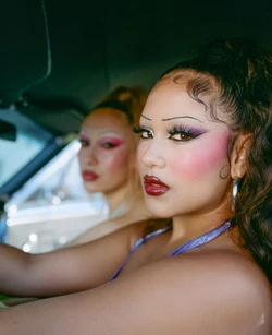 Two women with makeup sitting in a car bathed in sunlight, showcasing a blend of beauty and automotive elements.