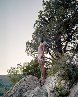 Shirtless man standing on rocky terrain outdoors with trees in the background under an overcast sky.