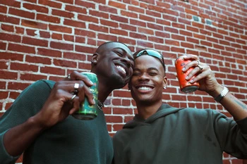 Two smiling Black men outdoors raise aluminum beverage cans in celebration against a textured red brick wall backdrop.