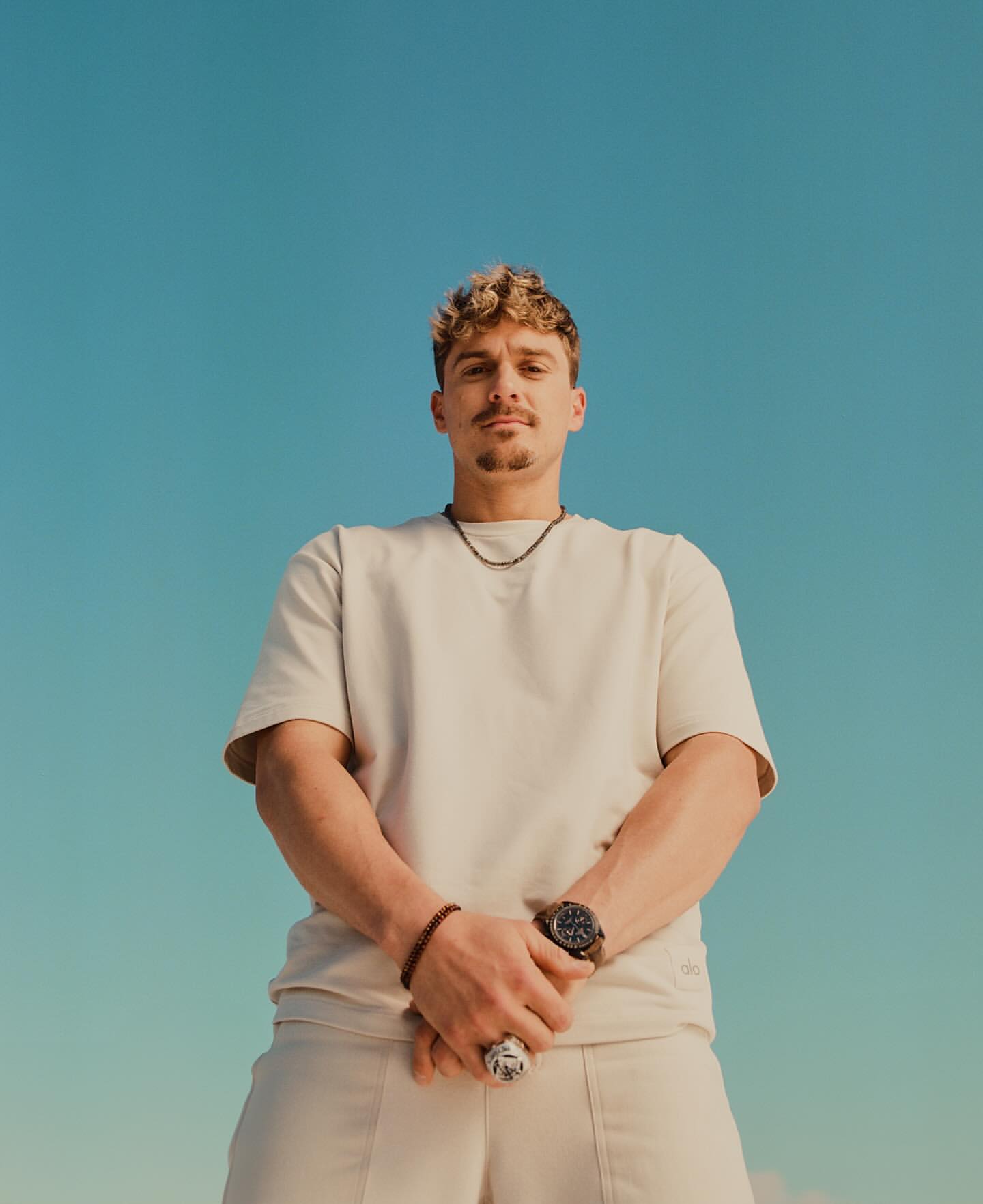 Low-angle shot of a light-skinned adult male with short, curly hair wearing a beige t-shirt and pants against a clear blue sky.