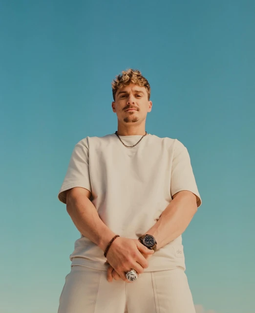 Low-angle shot of a light-skinned adult male with short, curly hair wearing a beige t-shirt and pants against a clear blue sky.