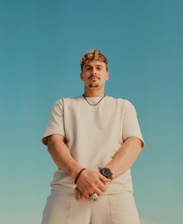 Low-angle shot of a light-skinned adult male with short, curly hair wearing a beige t-shirt and pants against a clear blue sky.
