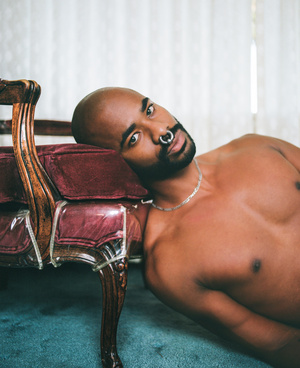 Portrait of a bald African American man with a nose ring, posing nude on a velvet chaise lounge, creating a sensual artistic photograph.