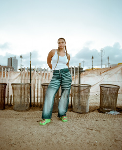 Young woman stands on a beach in jeans and a white top with a city skyline and sand dunes behind her, posing for a fashion editorial.