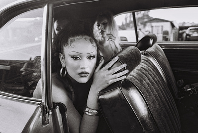 Black and white portrait inside a vintage car featuring a woman with dramatic makeup, hoop earrings, and two other women in the background.