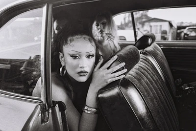 Black and white portrait inside a vintage car featuring a woman with dramatic makeup, hoop earrings, and two other women in the background.