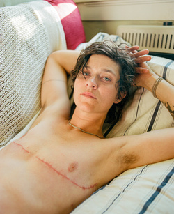 An adult female lies on a striped mattress with her hands behind her head, looking at the camera in natural light. 