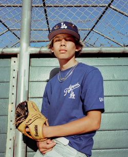 Teenage male in a Dodgers t-shirt and baseball cap holding a baseball glove outdoors in front of a chain-link fence on a sunny day.