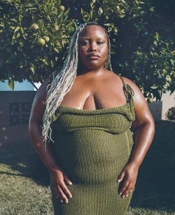 Portrait of a plus-size Black woman in an olive green dress standing outdoors with an orange tree in the background.