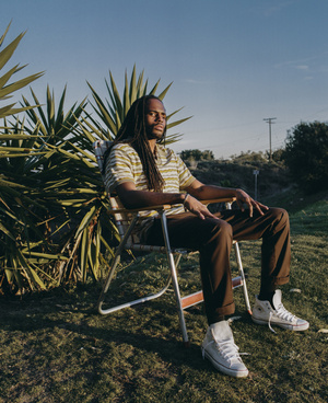 Portrait of a Black male model with long dreadlocks sitting on a lawn chair outdoors in a grassy field under a blue sky in the evening.