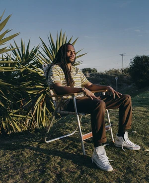 Portrait of a Black male model with long dreadlocks sitting on a lawn chair outdoors in a grassy field under a blue sky in the evening.