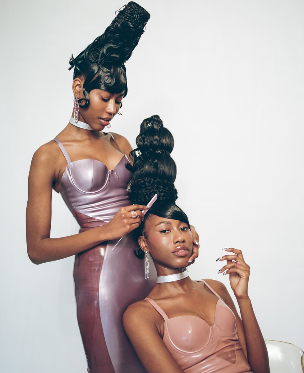 Two Black women in stylish dresses and elaborate hairstyles. One woman styles the other's hair in a studio setting with a white backdrop.