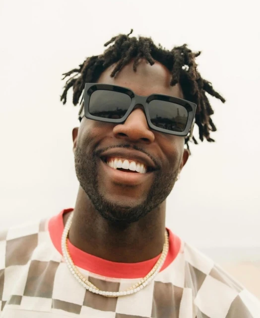 Smiling Black man in sunglasses and see-through checkerboard shirt against soft outdoor light. Close-up headshot with necklace.