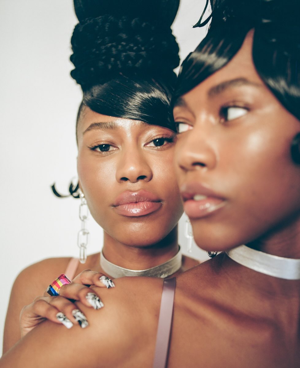Two African-descent women pose indoors. One has a high updo, silver choker and chain earrings. The other has short hair and white choker.
