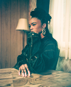Portrait of a black woman in a denim jacket with long pink nails seated at a wood grain table indoors