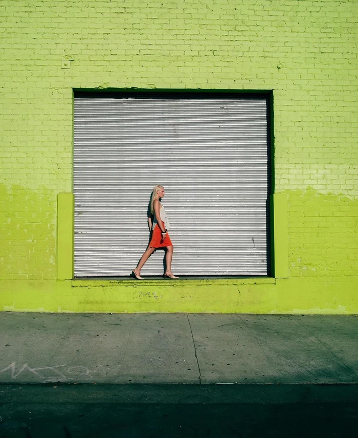 A blond woman walking past a large metallic roller shutter door set in a vibrant lime green brick wall.