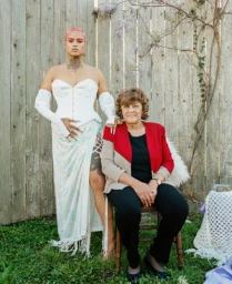Portrait of two women standing and seated together outdoors against a weathered wooden fence with vines