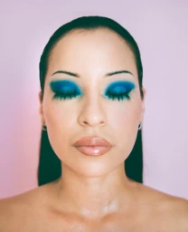 Close-up portrait of a woman with dramatic blue eyeshadow and long dark lashes against a pale pink background, photographed in Los Angeles.
