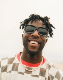 Portrait of a smiling Black man with dreadlocks wearing chunky black sunglasses and a patterned shirt, shot by Maiwenn Raoult.