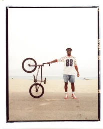 Man wearing shorts and patterned jersey holding up a black bicycle on a paved path near a beach.