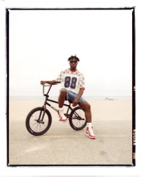 Black man in patterned jersey and denim shorts sitting on a BMX bicycle on a sandy beach by Maiwenn Raoult